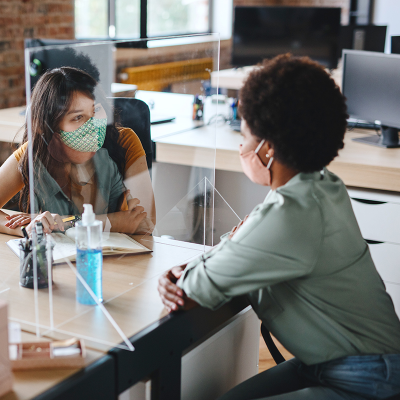 Two women wearing face masks sitting at a wooden table with a perspex screen between them