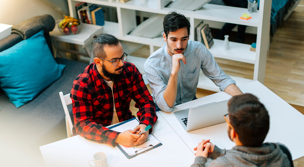 Three young men sitting at a table discussing ideas over a laptop
