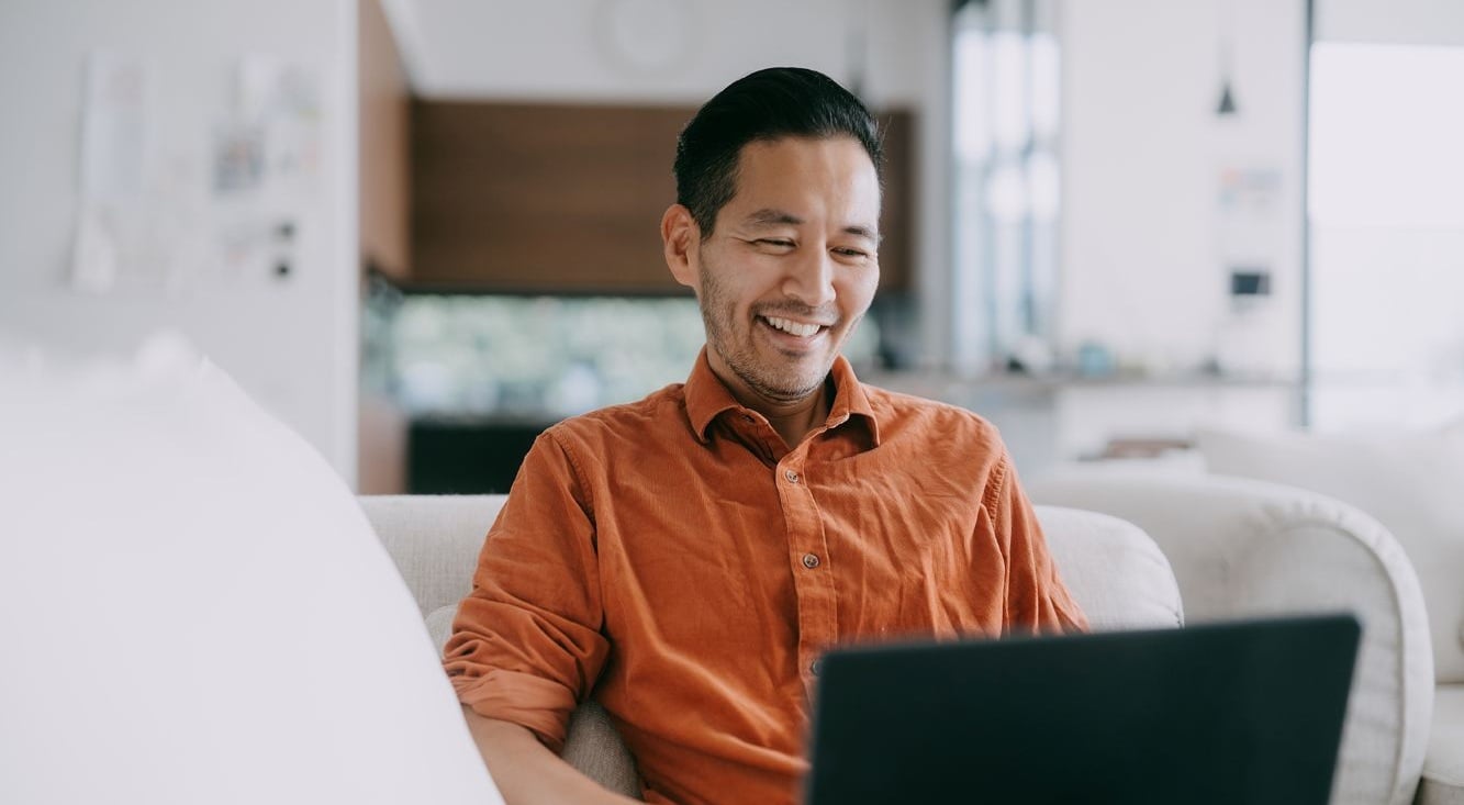 A man sitting on a light-coloured couch using a laptop in a bright, modern living room with soft natural light and minimalistic decor
