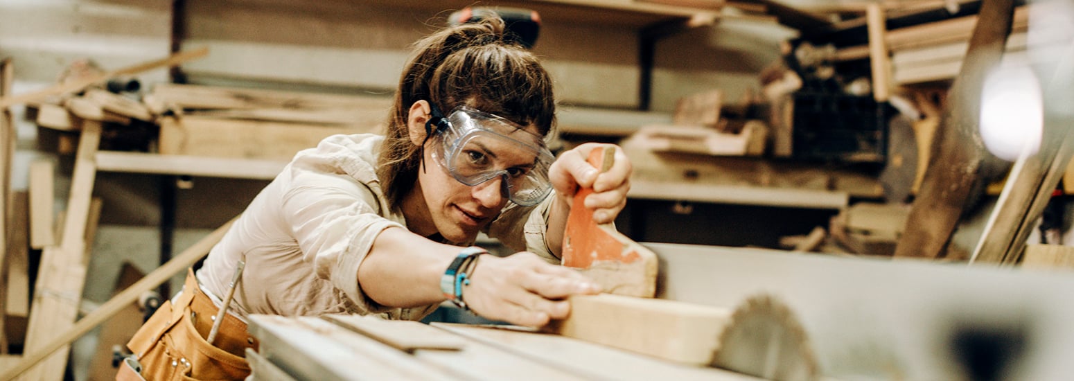 Young craftperson with protective equipment working with planks in workshop