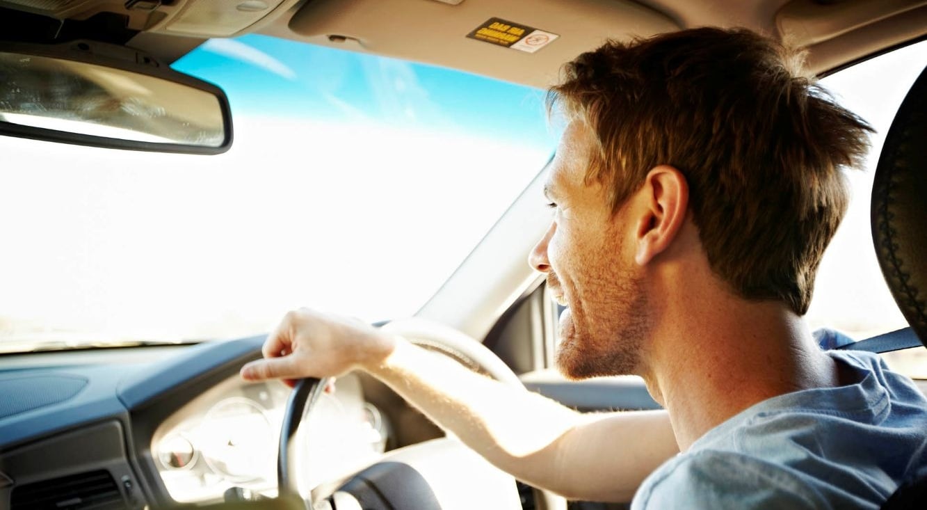 Interior view of a car with a man driving, one hand on the steering wheel and sunlight visible through the front windscreen.