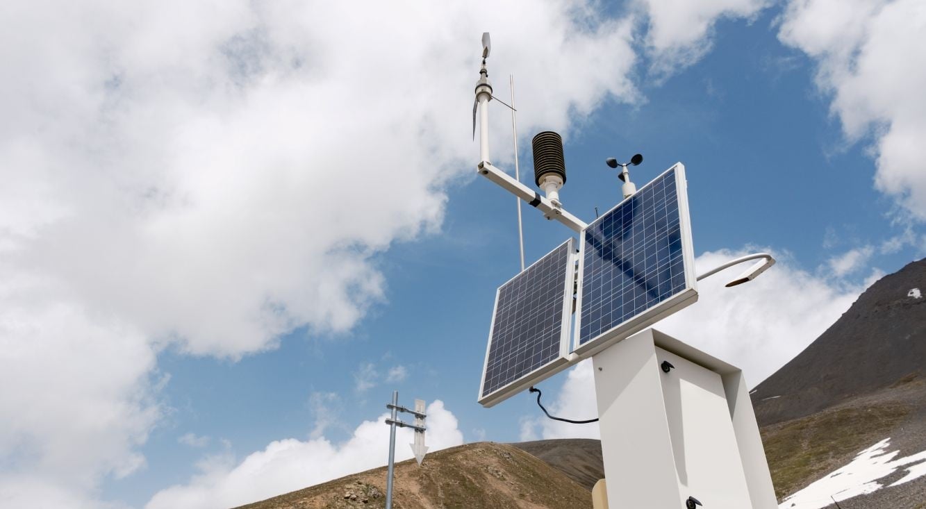 Remote weather monitoring station with solar panels and sensors installed in a mountainous landscape beneath a cloudy sky