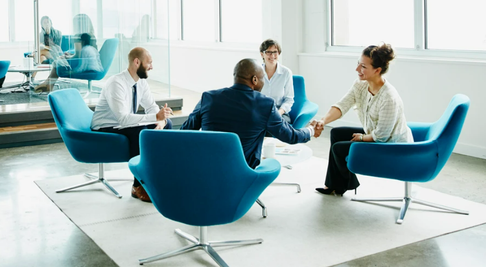 Four people meeting sitting in blue chairs, two people shake hands.