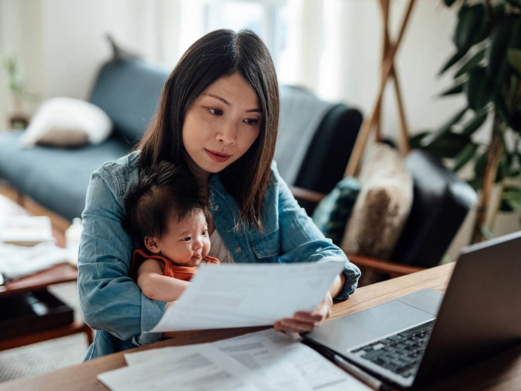 Young Asian mother reviewing her home insurance on her laptop while taking care of her baby sitting on her lap.