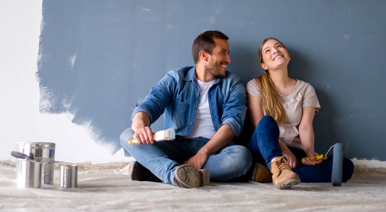 A couple sitting on a floor with paint brushes and rollers, painting an interior wall during a home renovation