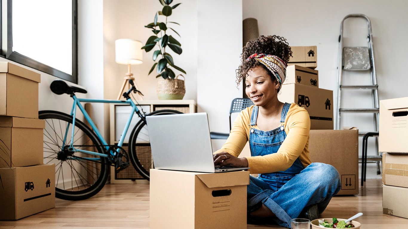 Woman using laptop while sitting on floor surrounded by moving boxes