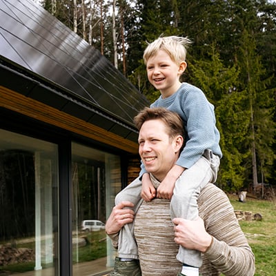 Father with young son on his shoulders in front of their family house with solar panels on the roof