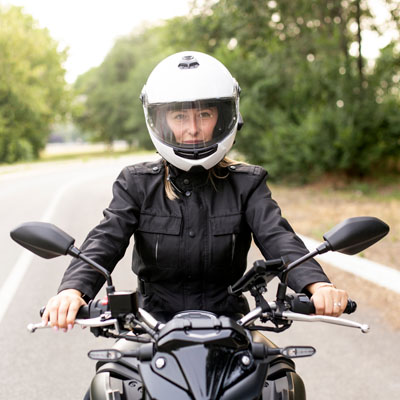 A female motorcyclist wearing a motorcycle helmet and protective leather jacket sitting on a bike on a tree-lined road, promoting QBE Comprehensive Motorcycle Insurance for Australian riders