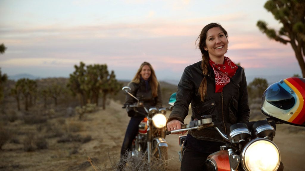 Two female motorcycle riders on a dirt track at sunset with helmets and bikes, promoting QBE Comprehensive Motorcycle Insurance for Australian riders