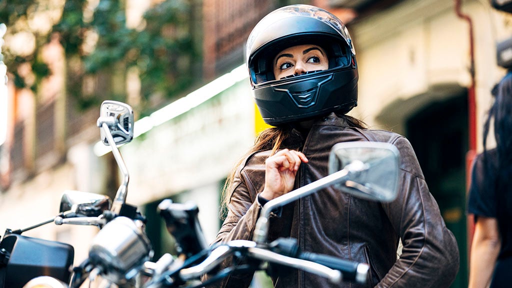 A female motorcyclist wearing a motorcycle helmet and riding gear while sitting on a motorcycle, preparing to ride on a city street.