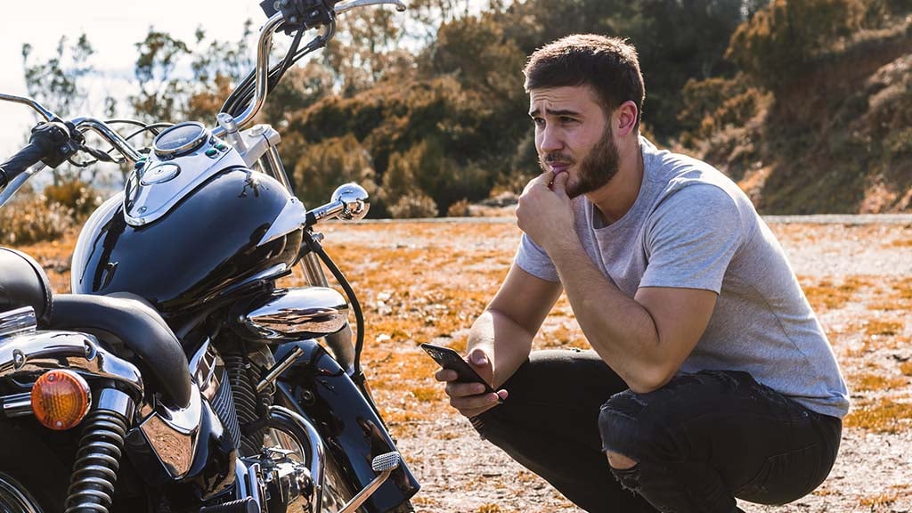 An anxious male motorcyclist crouches beside a parked black motorcycle on a scenic Australian roadside, holding his mobile phone – illustrating the steps to take after a motorbike accident also promote QBE motorcycle insurance.