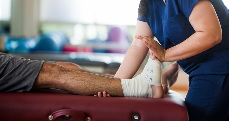 A healthcare professional supports an injured ankle during a rehabilitation session, with the worker lying on a treatment table in a clinical setting.
