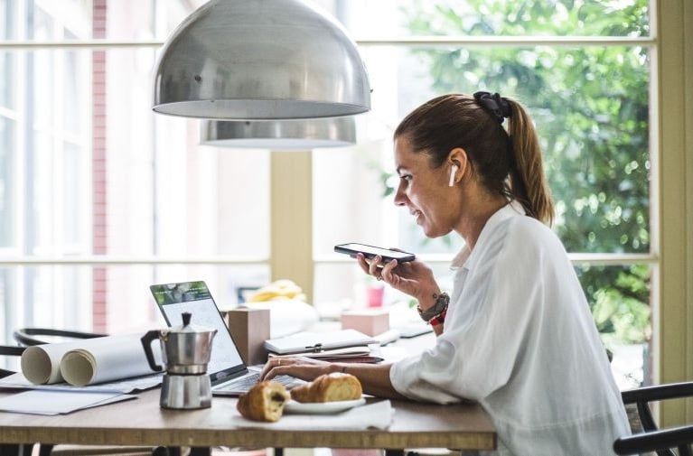 A woman seated at a desk in a bright home office, using a smartphone beside an open laptop, notebooks and a coffee maker, with pastries on the table and daylight coming through a window.