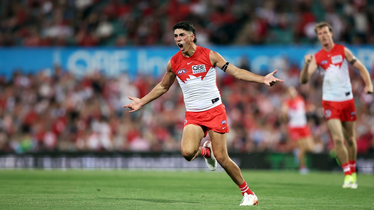 A Sydney Swans player in red and white running on the field with arms outstretched during an AFL match