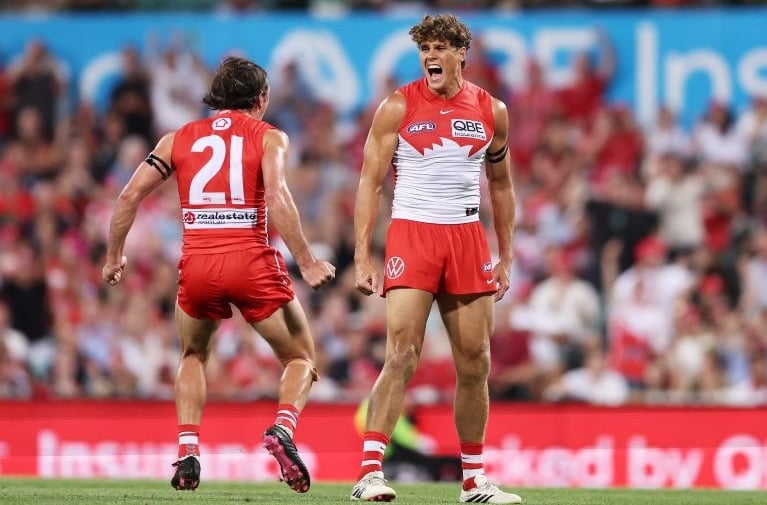 Sydney Swans AFL players on the field in red and white guernseys, with spectators visible in the stadium.