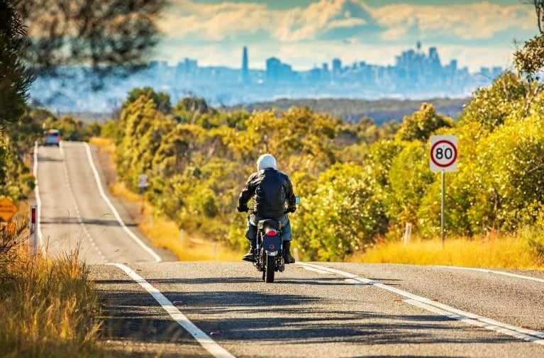 Image of a motorbike rider cruising down an open road with Sydney city skyline in the distance to promote comprehensive motorbike insurance in NSW, Australia.