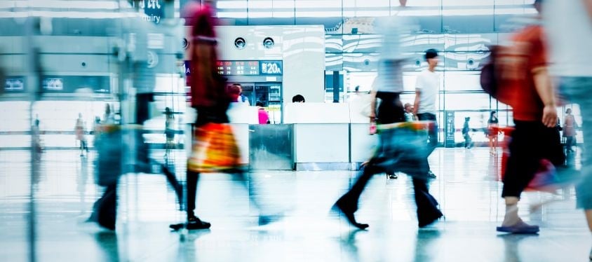Blurred travellers walk through a bright international airport terminal, with motion suggesting busy foot traffic, glass walls and departure signage visible in the background.