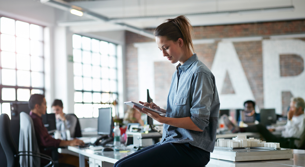 Woman sitting on her open-plan workplace desk, using a tablet