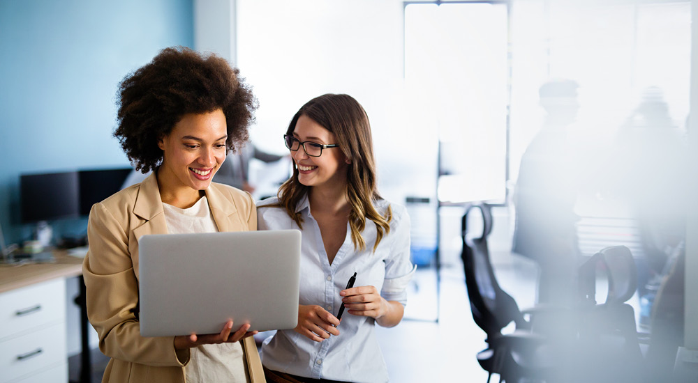 Two office workers stand side by side in a modern workspace, looking at a laptop held by one person while the other holds a pen