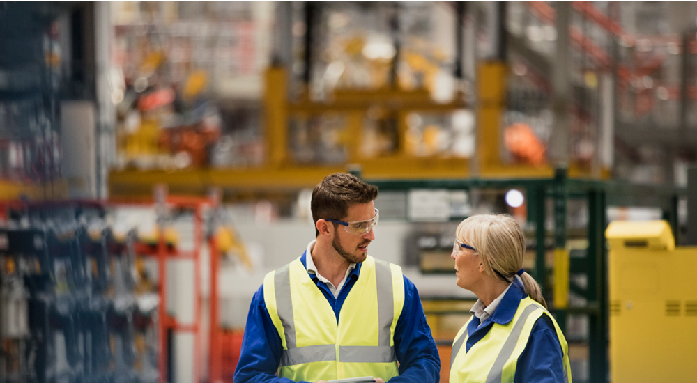 Two people wearing high‑visibility vests and safety glasses walking through a warehouse, with industrial shelving in the background.