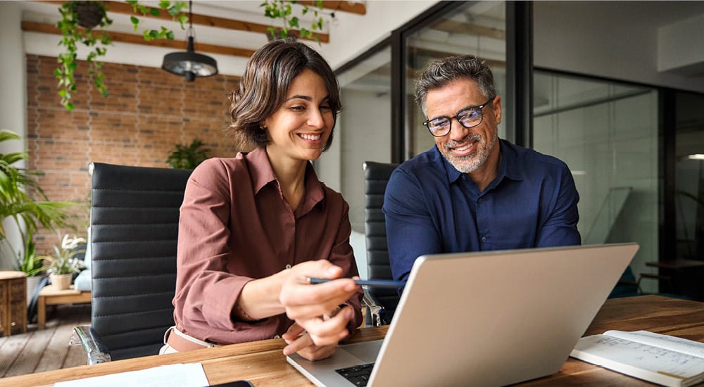 Two professionals collaborating at a desk, looking at a laptop and discussing information in a contemporary office setting