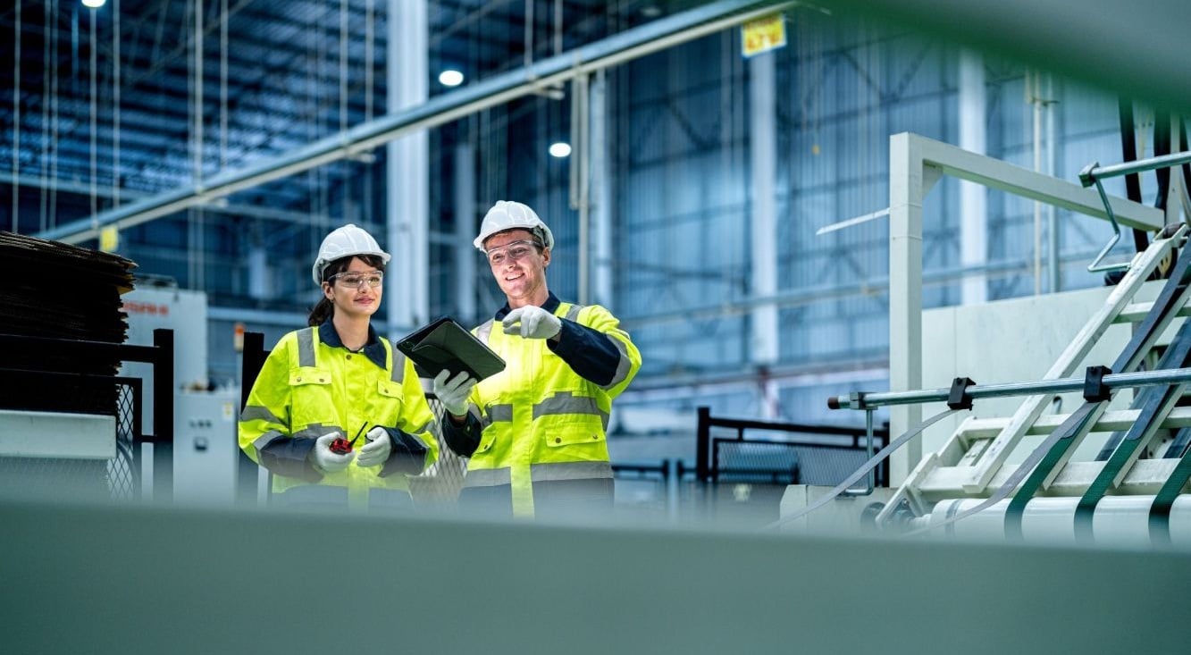 Two workers in a warehouse wearing high-vis gear reviewing information on a tablet beside industrial equipment