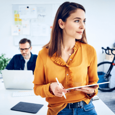 Woman leaning against a desk holding a tablet and stylus with colleague in the background