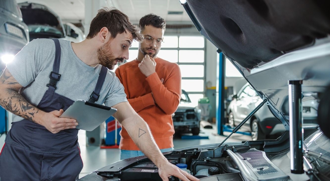 A mechanic inspecting a vehicle engine while discussing repairs with another person in an automotive workshop