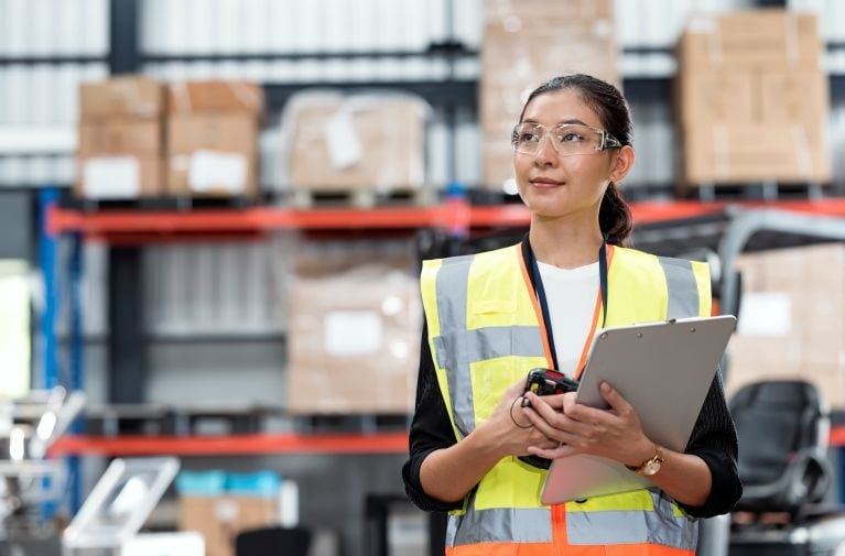Person wearing a high-visibility vest and safety glasses holding a clipboard in a warehouse with shelving and packaged goods in the background