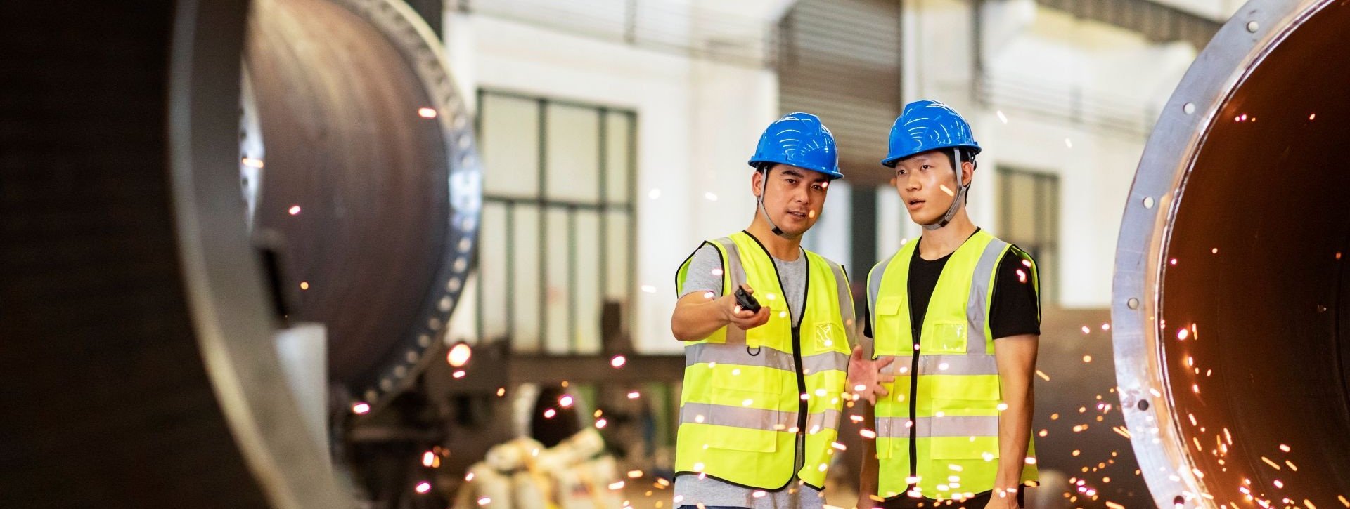 Two workers in safety helmets and high-visibility clothing inspect large steel pipes inside a manufacturing facility, with sparks visible from nearby metalworking activity