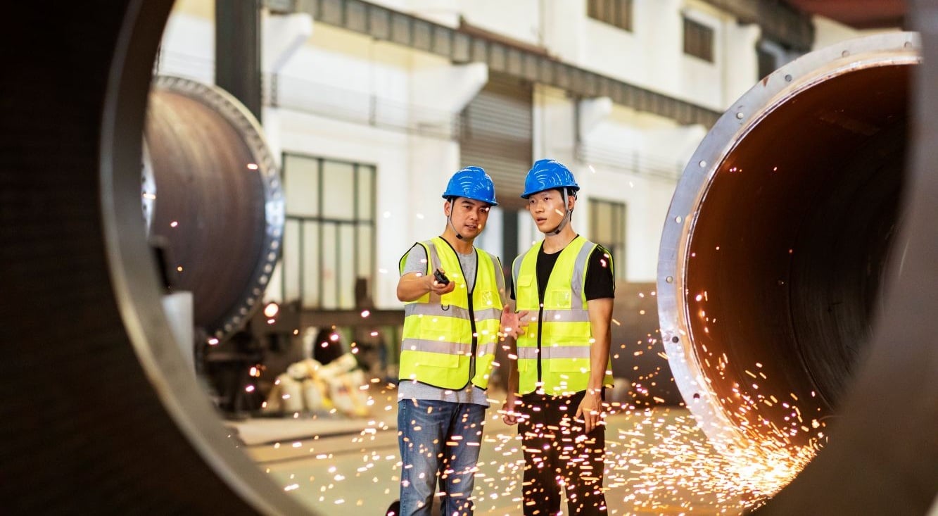 Two workers in safety helmets and high-visibility clothing inspect large steel pipes inside a manufacturing facility, with sparks visible from nearby metalworking activity