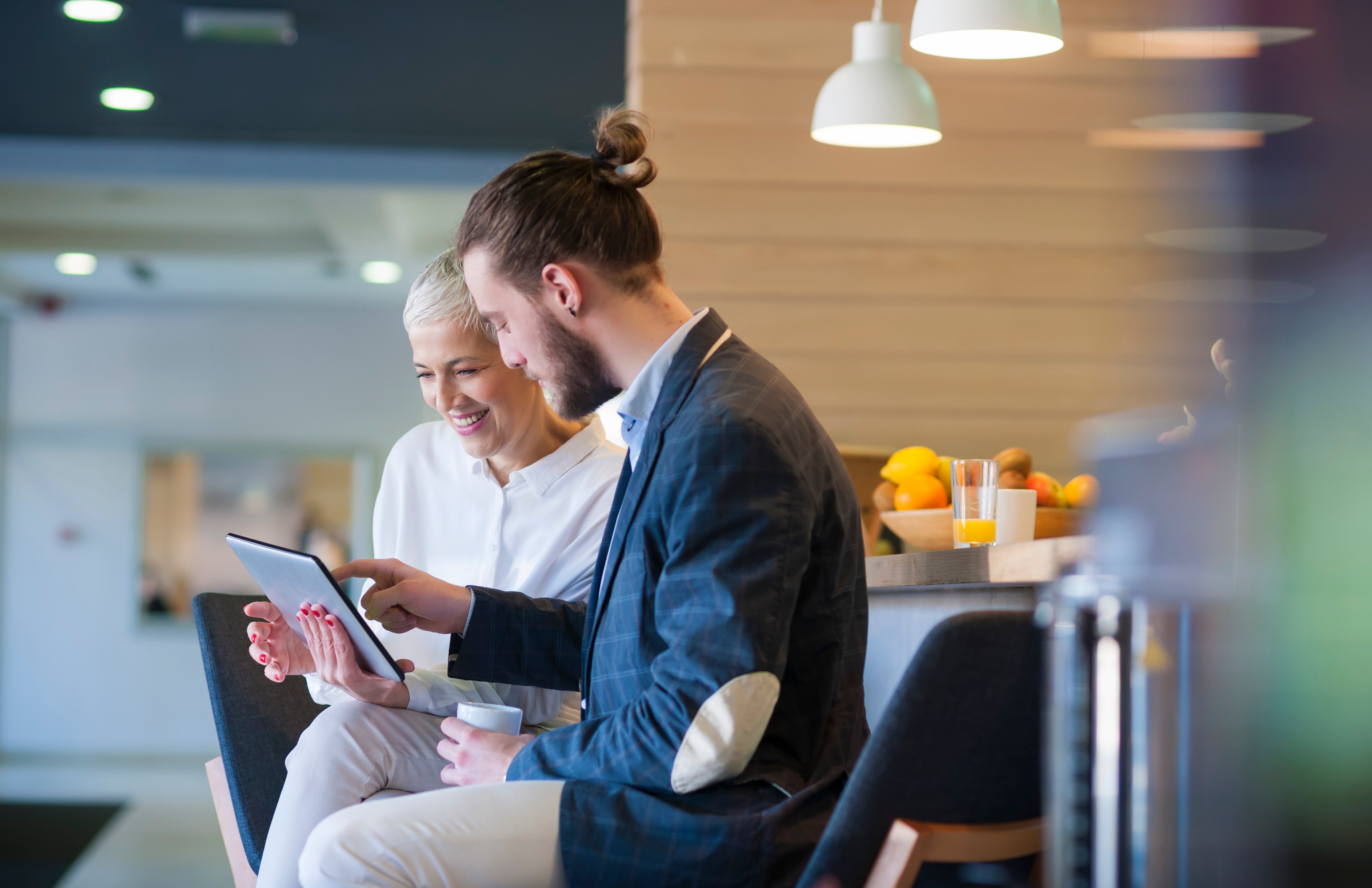 Two people sit side by side in a modern office kitchen area, looking at a tablet