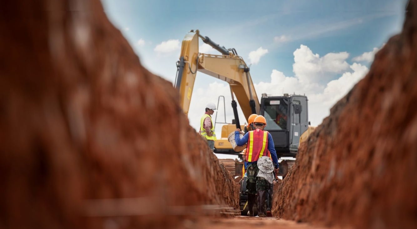 Two workers wearing safety equipment stand in a trench at a construction site, with an excavator positioned above them and earth embankments on either side
