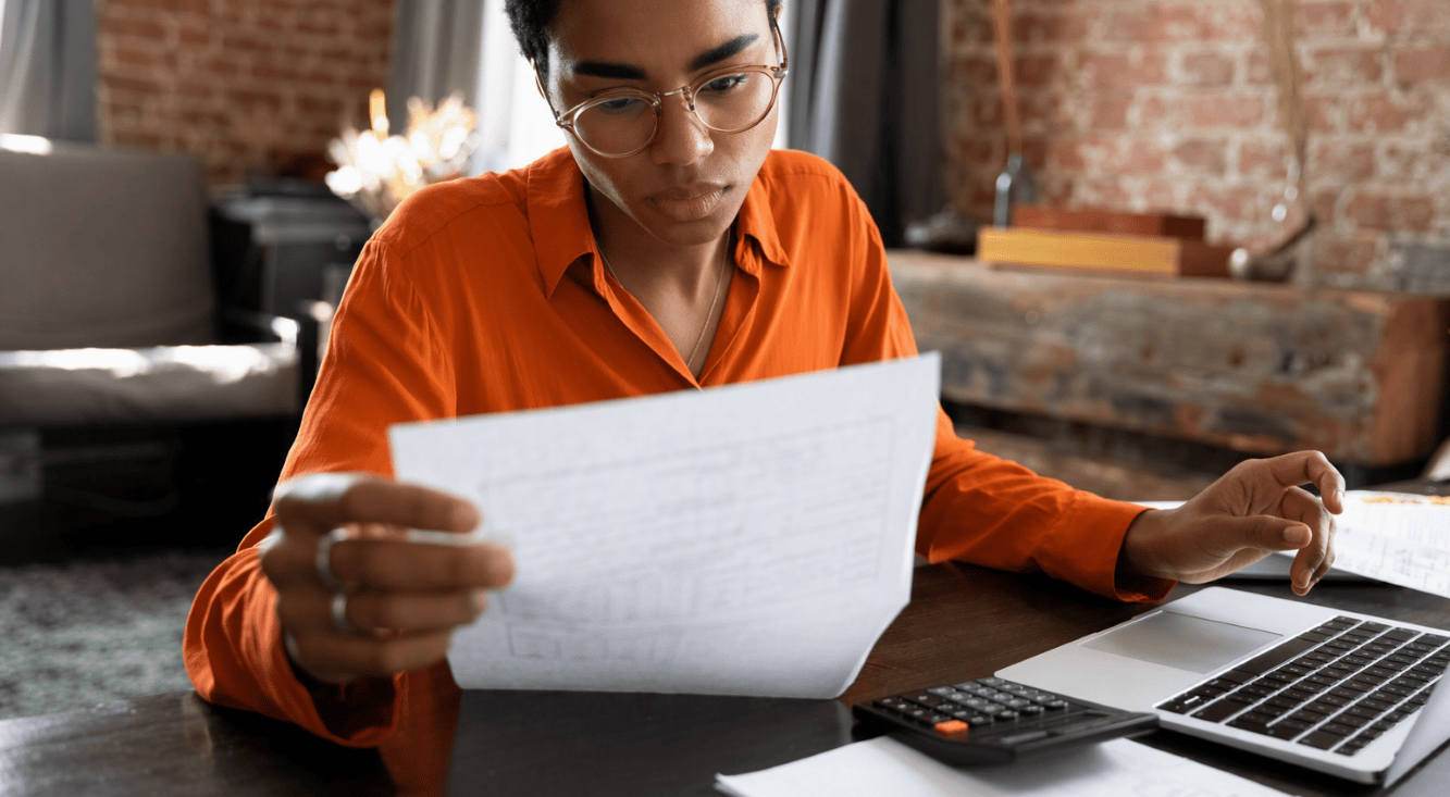 A woman reviewing paperwork at a desk with a laptop and calculator, wearing an orange shirt