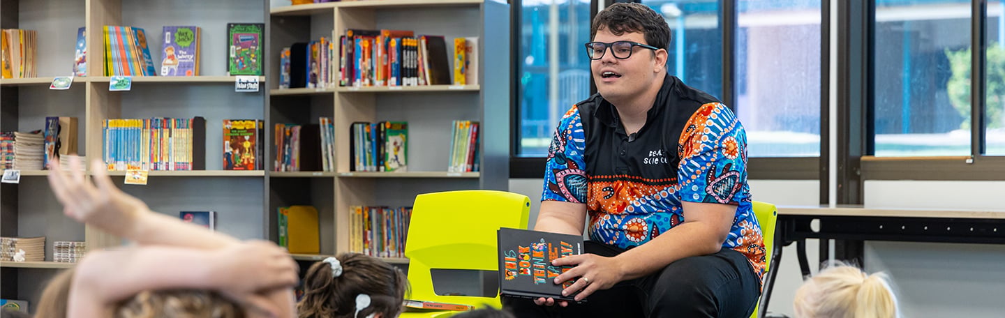 Person seated in a library holding a book while speaking to a group of children, with shelves of colourful books in the background