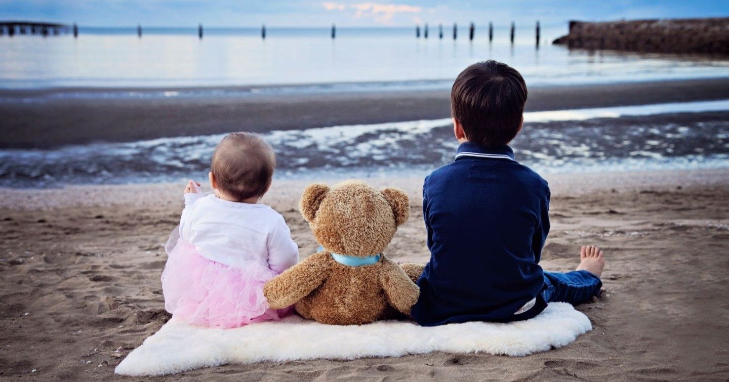 Two children and a teddy bear sitting on a beach blanket facing the water, symbolising remembrance and support for families grieving the loss of a child, as part of Precious Wings’ mission.