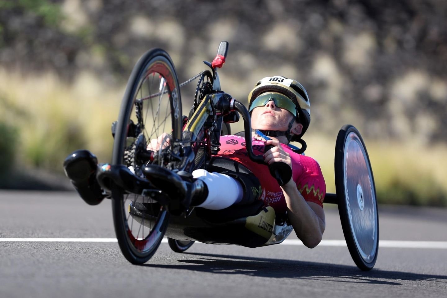 Australian Paralympian Lauren Parker propelling herself forward on an open road, wearing a pink racing suit, helmet numbered 103, and reflective sunglasses.