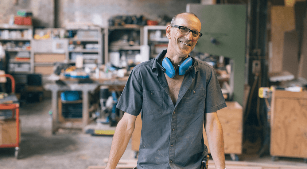 Mature man standing at a workbench in a warehouse with blue ear defenders around his neck