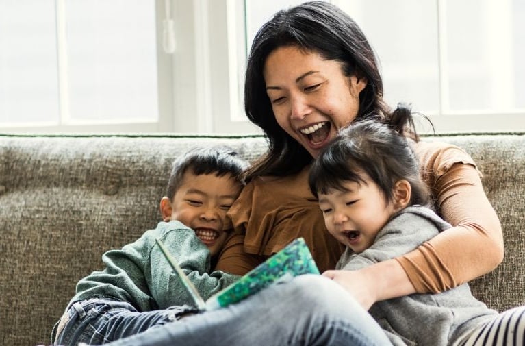 A mother and two children sitting together on a sofa, sharing a book in a living room.