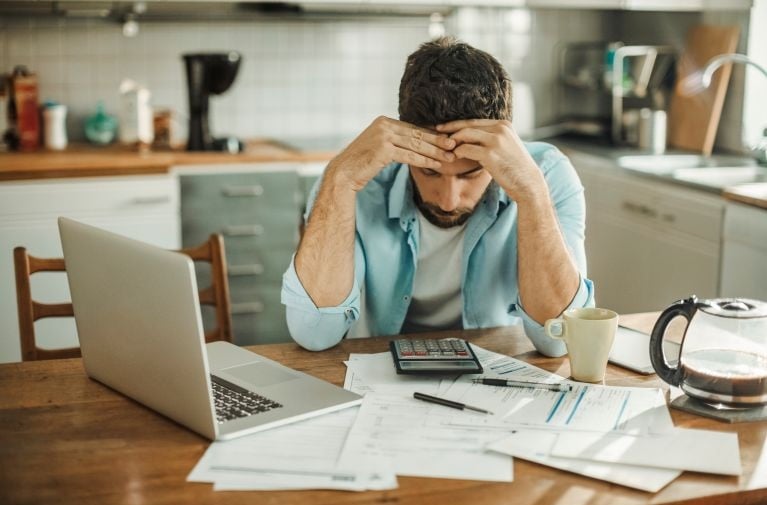 A man seated at a table with a laptop, paperwork, and bills, representing financial hardship and difficulty meeting home loan repayments.