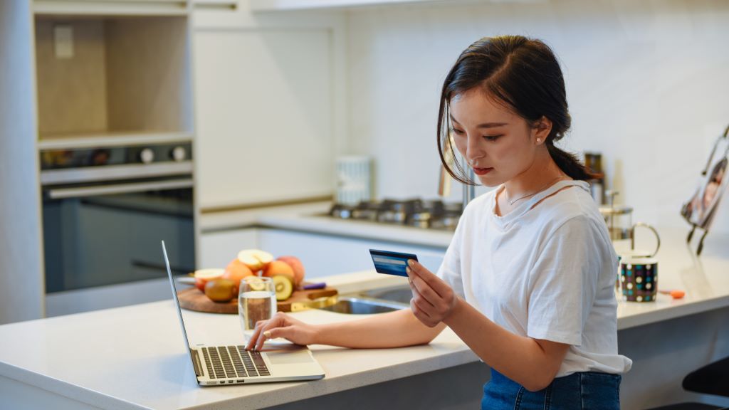 Young asian woman seated at kitchen counter looking at credit card while using laptop computer