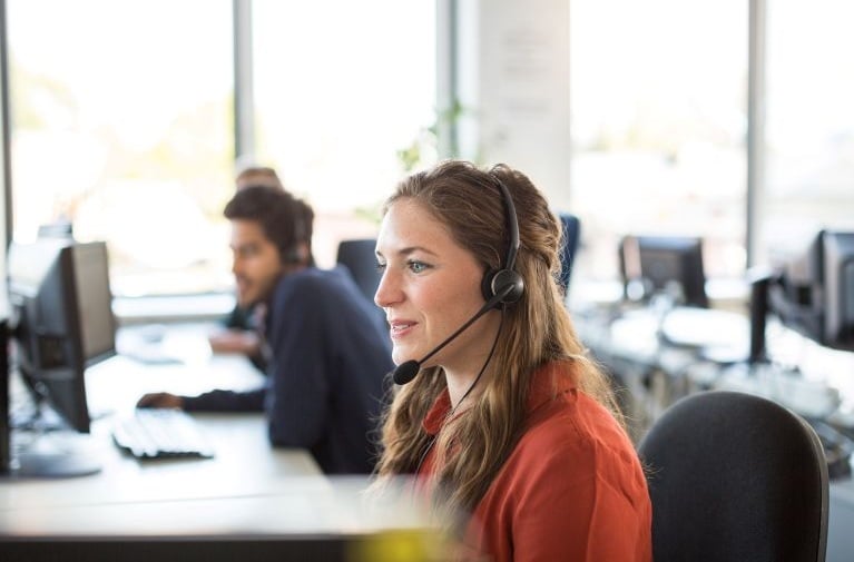 Customer service staff member wearing a headset, seated at a desk in a modern office with computers in the background