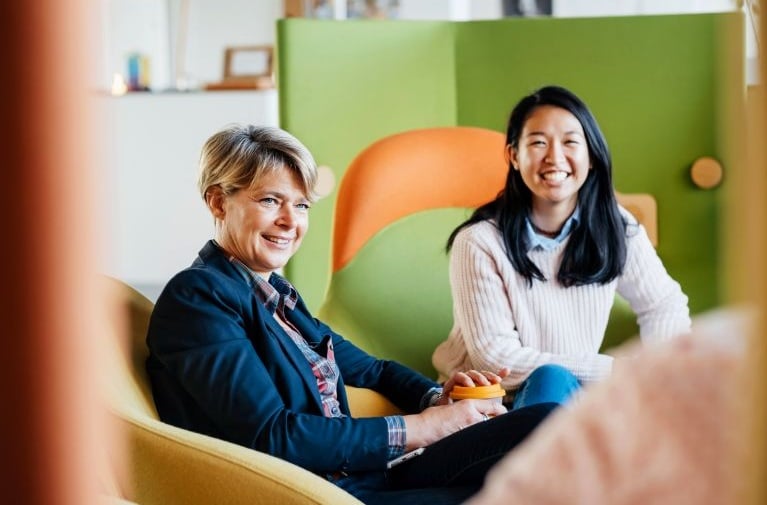 Two colleagues seated in a casual meeting area, holding takeaway coffee cups, with colourful furniture in the background