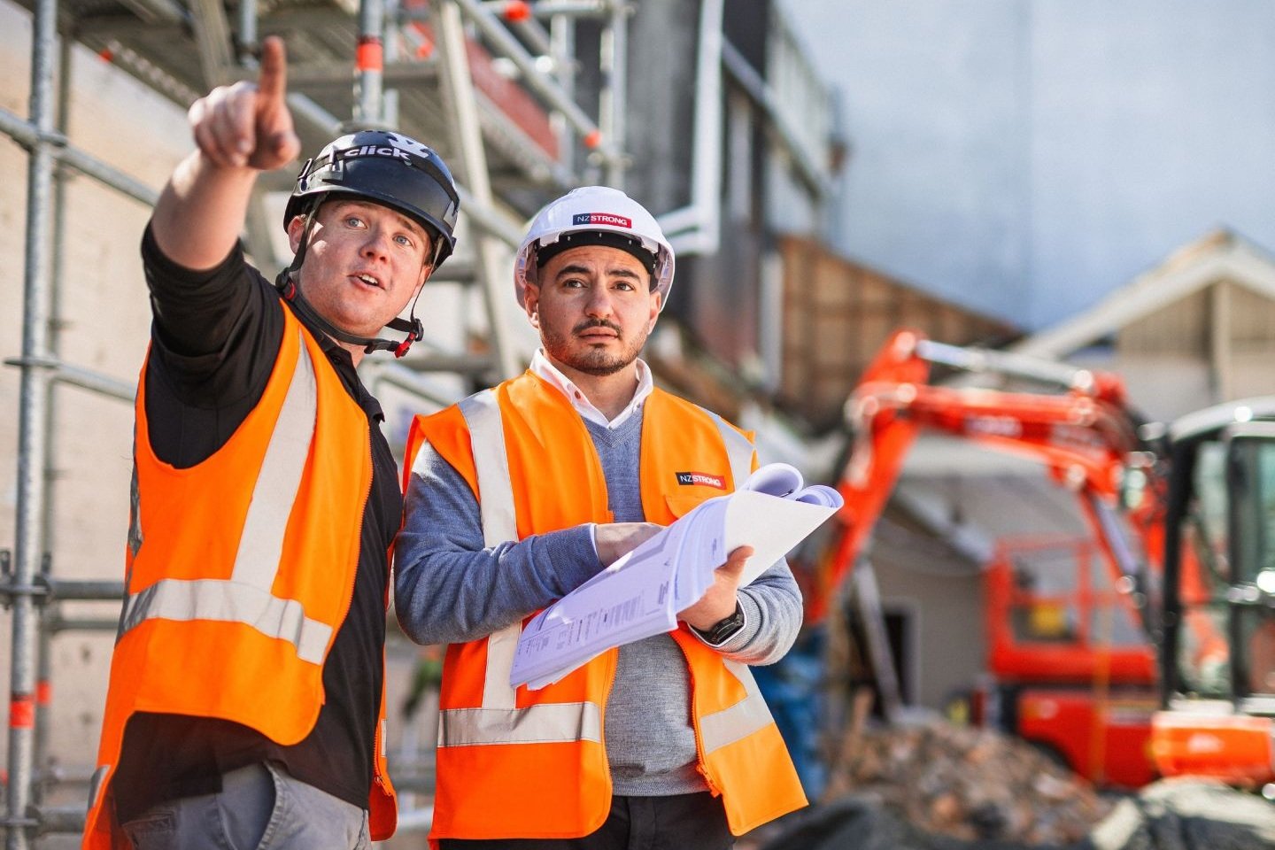 Two construction workers wearing high-visibility orange safety vests and hard hats stand at a building site. One worker is pointing towards the structure while the other holds open building plans. Scaffolding and an orange excavator are visible in the background, indicating active construction work.