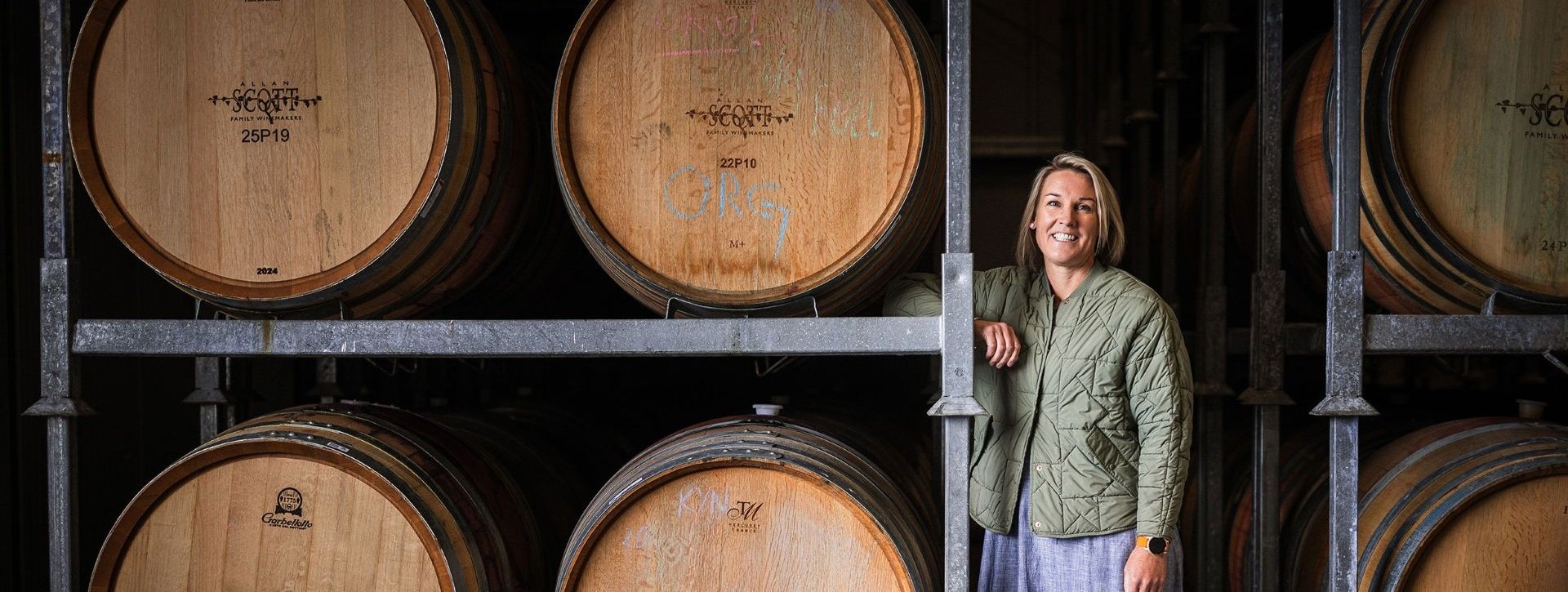 A woman standing beside stacked wooden barrels inside a winery storage area